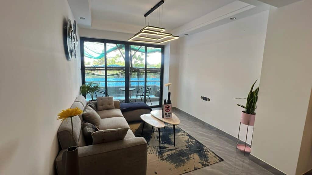 Contemporary living room lounge featuring grey sectional sofa, geometric pendant lighting, and sliding glass doors to the balcony at Serenity Residence in Kilimani, Nairobi.