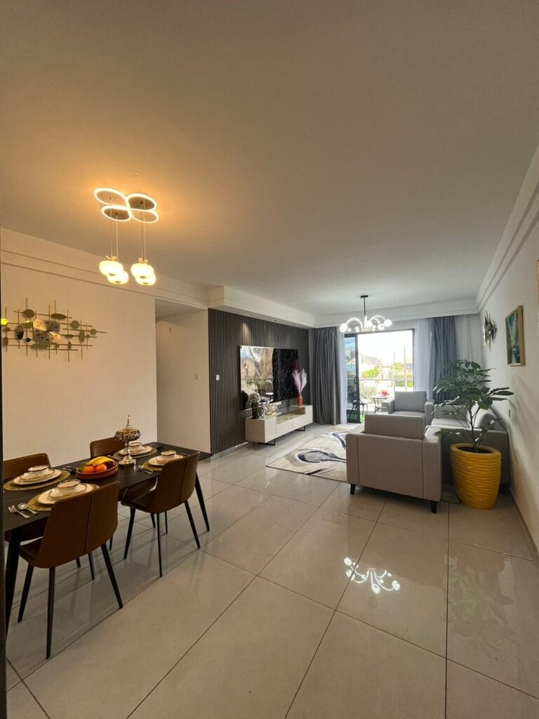 Spacious open-plan living room and dining area with fluted TV accent wall, modern ceiling light fixtures, and balcony access at New Rise Gardens in Syokimau, Nairobi.