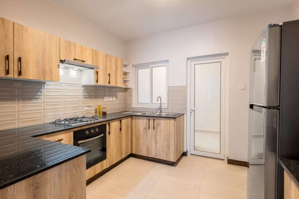 L-shaped fitted kitchen with warm wood-grain cabinetry, dark granite countertops, built-in gas stove, oven, and utility door access at Sunpark Residences in Syokimau, Nairobi.